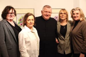 Portret grupowy pięciu uśmiechniętych osób stojących blisko siebie na tle jasnej ściany galerii. Od lewej: kobieta w okularach i szarym żakiecie, kobieta w białej koszuli z ozdobnym detalem, mężczyzna w czarnej sutannie (ks. Tadeusz Pajurek), kobieta w okularach i marynarce w drobną kratkę oraz kobieta w brązowym kardiganie (Barbara Maślak). W tle, między postaciami, widoczne są fragmenty oprawionych prac plastycznych przedstawiających kolorowe kwiaty.