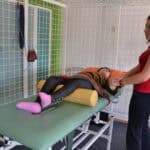 Zajęcia rehabilitacyjne. Osoba leży na łóżku rehabilitacyjnym z wałkiem pod kolanami i unosi lekko stopę. Obok łóżka stoi kobieta i trzyma jej rękę
