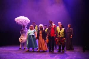 Grupa aktorów w barwnych kostiumach stoi w rzędzie na scenie, kłania się widowni. Na pierwszym planie widać kobietę w jasnoniebieskiej sukni i diademie, trzymającą za rękę inną aktorkę w futrzanej czapce i długiej spódnicy. Obok nich stoi mężczyzna w czerwono-szarym stroju z godłem. W tle po lewej postać w kostiumie bałwana trzyma dużą, białą, pierzastą parasolkę, a pozostali aktorzy mają na sobie m.in. zielone kamizelki imitujące łuski. Scena oświetlona jest fioletową łuną