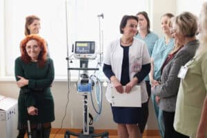Siedem kobiet stoi na jasnym korytarzu szpitalnym obok respiratora Hamilton Medical. Renata Polisiakiewicz stoi po lewej stronie, ma skrzyżowane ręce i trzyma kulę. W centrum stoi lekarka w białym fartuchu (Renata Nieznaj) trzyma białą teczkę. Pozostałe kobiety, ubrane w stroje wizytowe i mundurki medyczne, stoją po obu stronach grupy, patrząc w stronę obiektywu lub na siebie nawzajem.