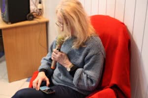Blondynka w niebieskim, plecionym swetrze siedzi na fotelu z czerwonym kocem. W jednej ręce trzyma złoty mikrofon blisko ust, a drugą ręką obsługuje smartfon leżący na jej kolanie, z którego czyta tekst. Na ręku ma złoty zegarek. W tle widoczny jest fragment drewnianego biurka ze sprzętem elektronicznym i kablami