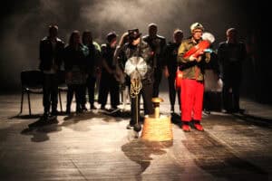 Scena z przedstawienia teatralnego z udziałem grupy aktorów w kostiumach nawiązujących do różnych zawodów. Na pierwszym planie po prawej stoi mężczyzna w hełmie i czerwonych spodniach, trzymający zwinięty wąż strażacki. Obok niego, w centrum, aktor w skórzanej kurtce i cylindrze prezentuje rekwizyt przypominający kominiarską szczotkę nad makietą komina. W tle widać pozostałych wykonawców, m.in. osobę w mundurze policyjnym oraz w kurtkach motocyklowych. Scena jest spowita dymem i oświetlona ciepłym światłem reflektorów