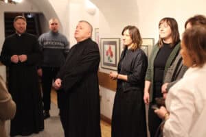 Grupa osób stoi w korytarzu galerii, patrząc w jednym kierunku podczas części oficjalnej wydarzenia. Na pierwszym planie widać starszego duchownego w czarnej sutannie ze splecionymi dłońmi, a obok niego kobietę w czarnym stroju i drugą w zielonym żakiecie. W tle, po lewej stronie, stoi drugi ksiądz (ks. Bogusław Suszyło) oraz mężczyzna w niebieskim golfie. Ściany pomieszczenia zdobią oprawione grafiki, a w głębi widoczne są schody prowadzące na wyższe piętro.