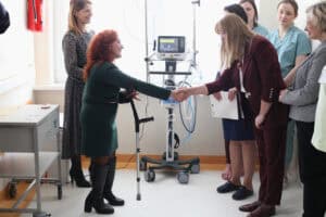 Moment uścisku dłoni pomiędzy Renatą Polisiakiewicz a Ewą Mańdziuk. Obie kobiety są uśmiechnięte i lekko pochylone ku sobie. W tle stoją pozostałe uczestniczki spotkania oraz respirator medyczny.