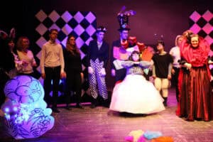 Scena zbiorowa z przedstawienia inspirowanego „Alicją w Krainie Czarów”. Na pierwszym planie po prawej stoi aktorka w bogato zdobionej, czerwono-czarnej sukni Królowej Kier, a w centrum postać w szerokiej białej spódnicy. W tle widoczni są inni aktorzy, m.in. Szalony Kapelusznik w wysokim cylindrze oraz osoby w strojach z motywem czarno-białej szachownicy. Po lewej stronie znajduje się duża, świecąca dekoracja w kształcie kwiatu. Scena oświetlona jest różowym światłem, a na podłodze leżą kolorowe kawałki materiału