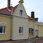 Budowa Zakładu Aktywności Zawodowej przy SION w Bartoszycach. Parterowy budynek o żółtej elewacji, przy dwóch oknach stoją drabiny. Na jednej drabinie mężczyzna wykonuje obróbki stolarki okiennej