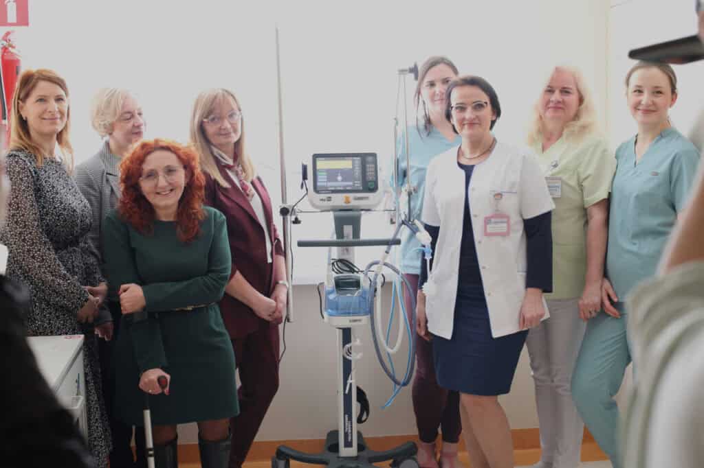 Osiem kobiet pozuje do zdjęcia w jasnej przestrzeni szpitalnej. Wszystkie są zwrócone w stronę obiektywu i uśmiechają się. W centralnym punkcie kompozycji znajduje się respirator Hamilton Medical. Grupa składa się z kobiet w strojach biznesowych, codziennych oraz personelu medycznego w fartuchach i mundurkach