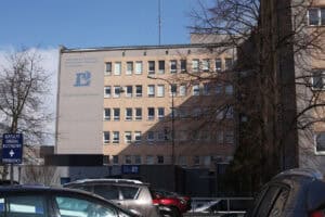 Wielopiętrowy budynek Samodzielnego Publicznego Zakładu Opieki Zdrowotnej w Lubartowie. Na elewacji widać duże niebieskie logo z literą „L” i symbolem medycznym oraz adres: ul. Cicha 14, 21-100 Lubartów. Po lewej stronie znajduje się niebieska tablica z napisem: Szpitalny Oddział Ratunkowy + Emergency. Na pierwszym planie zaparkowane samochody i bezlistne drzewa