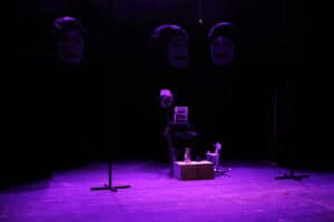 Mroczna scena teatralna w fioletowym świetle. Trzy wysokie, czarne bryły z maskami są rozstawione w tle. Na środku sceny siedzi postać, która zamiast maski ma na głowie białą klatkę dla ptaków. Obok niej znajdują się białe rekwizyty: mały koń na biegunach, kufer oraz lampa naftowa. Postać trzyma długi kij zwieńczony białym kapeluszem. Cała kompozycja jest symetryczna, z czarnymi statywami po bokach, co potęguje surrealistyczny nastrój przedstawienia