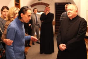 Na pierwszym planie, po prawej stronie, stoi starszy duchowny (ks. Tadeusz Pajurek) w czarnej sutannie ze splecionymi dłońmi, patrząc łagodnie przed siebie. Po lewej stronie widoczna jest kobieta (Alicja Czajka) w niebieskim, wzorzystym golfie, którą za ramię podtrzymuje inna osoba. W tle, w głębi jasnego korytarza o łukowych sklepieniach, stoi grupa uczestników wydarzenia, w tym mężczyzna w szarym garniturze oraz drugi duchowny. Sceneria obejmuje fragmenty drewnianych ław oraz schody prowadzące na wyższe piętro galerii.