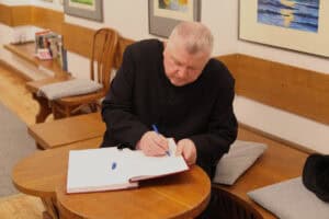 Duchowny (ks. Tadeusz Pajurek) w czarnej sutannie siedzi na niskiej, drewnianej ławie z szarymi poduszkami i wpisuje się do otwartej księgi pamiątkowej leżącej na małym, okrągłym stoliku. W prawej dłoni trzyma niebieski długopis, a obok, na białej kartce, leży skuwka. W tle widoczny jest korytarz galerii z jasnymi ścianami, na których wyeksponowano oprawione grafiki. Za plecami mężczyzny, na drewnianym regale, ustawionych jest kilka książek, a obok stoi puste drewniane krzesło z szczebelkowym oparciem.