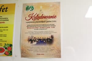 Zbliżenie na plakat informacyjny o treści: „Kolędowanie z pacjentami, pracownikami i gośćmi COZL”. Plakat zaprasza na koncert kolęd w Centrum Onkologii Ziemi Lubelskiej, który odbył się 28 stycznia 2026 r. o godz. 16:00. Na plakacie widnieje zdjęcie chóru LORIEN pod kierownictwem Katarzyny Kulszew oraz informacja o prowadzącej – prof. Iwonie Kalinowskiej. W tle plakatu motyw świąteczny z gwiazdą i nutami, a obok fragment innego plakatu.