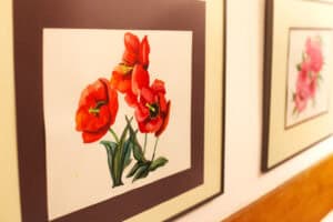 Bliskie ujęcie dwóch oprawionych grafik wiszących na białej ścianie galerii. Na pierwszym planie widoczna jest praca przedstawiająca trzy czerwone maki z zielonymi łodygami, ujęte w ciemne passe-partout i jasną ramkę. W tle, nieco nieostre, widoczne jest drugie dzieło przedstawiające bukiet różowych kwiatów. Poniżej obrazów przebiega fragment poziomej, drewnianej listwy wykończeniowej.