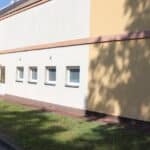 Wejście do parterowego budynku Sali rehabilitacyjnej przy SION w Bartoszycach. Biało-beżowa elewacja, drzwi i cztery małe okienka. Przed budynkiem zielony trawnik, budynek częściowo w słońcu, na elewacji cienie drzewa, w tle kawałek błękitnego nieba