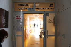 Widok przez otwarte przeszklone drzwi na jasny korytarz Oddziału Położniczo-Noworodkowego. Nad wejściem widnieje duży napis „ODDZIAŁ POŁOŻNICZO - NOWORODKOWY” oraz logo szpitala. Na szybie drzwi umieszczono piktogram przedstawiający kobietę karmiącą niemowlę. W głębi korytarza widać sylwetki personelu medycznego na tle silnego światła padającego z końca przejścia. Po lewej stronie na ścianie wisi fotografia noworodka