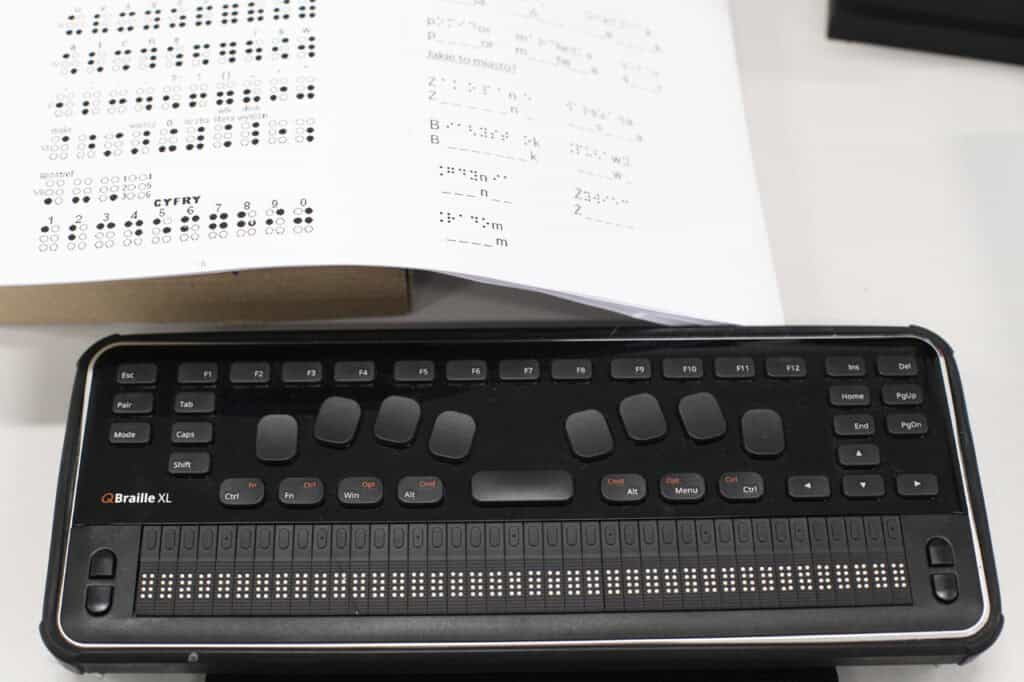 Specjalistyczny terminal brajlowski QBraille XL leży na jasnym biurku. Urządzenie jest czarne, ma prostokątny kształt i łączy funkcje klawiatury komputerowej z monitorem brajlowskim. W centralnej części umieszczono osiem dużych, ergonomicznych klawiszy do pisania brajlem oraz szeroką spację. Dolną część urządzenia zajmuje długi pasek z czterdziestoma aktywnymi komórkami brajlowskimi, które umożliwiają dotykowy odczyt tekstu. W tle, nad urządzeniem fragment białej karty z wydrukowanym alfabetem Braille’a oraz ćwiczeniami językowymi.