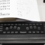 Specjalistyczny terminal brajlowski QBraille XL leży na jasnym biurku. Urządzenie jest czarne, ma prostokątny kształt i łączy funkcje klawiatury komputerowej z monitorem brajlowskim. W centralnej części umieszczono osiem dużych, ergonomicznych klawiszy do pisania brajlem oraz szeroką spację. Dolną część urządzenia zajmuje długi pasek z czterdziestoma aktywnymi komórkami brajlowskimi, które umożliwiają dotykowy odczyt tekstu. W tle, nad urządzeniem fragment białej karty z wydrukowanym alfabetem Braille’a oraz ćwiczeniami językowymi.