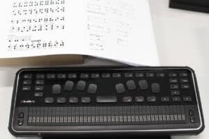 Specjalistyczny terminal brajlowski QBraille XL leży na jasnym biurku. Urządzenie jest czarne, ma prostokątny kształt i łączy funkcje klawiatury komputerowej z monitorem brajlowskim. W centralnej części umieszczono osiem dużych, ergonomicznych klawiszy do pisania brajlem oraz szeroką spację. Dolną część urządzenia zajmuje długi pasek z czterdziestoma aktywnymi komórkami brajlowskimi, które umożliwiają dotykowy odczyt tekstu. W tle, nad urządzeniem fragment białej karty z wydrukowanym alfabetem Braille’a oraz ćwiczeniami językowymi.
