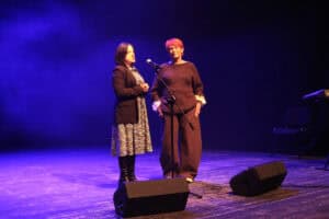 Dwie kobiety stoją na scenie przed mikrofonem na statywie. Kobieta po lewej, w ciemnej marynarce i wzorzystej sukience, mówi do mikrofonu, trzymając splecione dłonie. Kobieta po prawej (Maria Pietrusza-Budzyńska), o rudych włosach, ubrana w ciemny sweter i szerokie spodnie, patrzy przed siebie z lekkim uśmiechem. Scena jest oświetlona niebieskim światłem, w tle widać dym (efekt sceniczny), a na podłodze leżą dwa czarne odsłuchy sceniczne