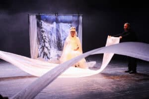 Scena teatralna o tematyce zimowej. W centrum, przed ekranem z grafiką ośnieżonych gór i lasu, siedzi kobieta w białej sukni, pelerynie i futrzanej czapce z diademem. Dwóch mężczyzn po bokach (jeden widoczny po prawej w czarnym stroju) trzyma i unosi długie pasy białego, półprzezroczystego materiału, imitujące zaspy śnieżne lub wiatr. Scena jest spowita dymem i oświetlona jasnym, chłodnym światłem, które podkreśla dynamikę ruchu tkaniny