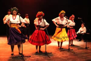 Grupa tancerzy w strojach ludowych występuje na scenie oświetlonej ciepłym, pomarańczowym światłem. Na pierwszym planie cztery kobiety w białych bluzkach, czerwonych koralach i kolorowych, rozłożystych spódnicach (fioletowej, czerwonej, żółtej i różowej) z kwiatowymi wiankami na głowach, pozują przy mikrofonach na statywach. W tle widać trzech mężczyzn w białych koszulach i ciemnych kamizelkach, a po prawej stronie siedzi chłopak grający na instrumencie perkusyjnym. Całość emanuje radosną, folkową energią