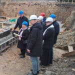 Nadanie aktu erekcyjnego dla Wspomaganej Społeczności Mieszkaniowej. Na placu budowy stoi kobieta i sześciu mężczyzn, w tym jeden w sutannie. Kobieta trzyma w jednej ręce połyskującą tubę na akt erekcyjny, a w drugiej kielnię