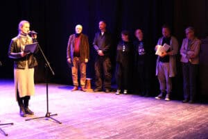 Na oświetlonej fioletowym światłem scenie stoi grupa osób podczas oficjalnej części wydarzenia. Po lewej stronie, przy mikrofonie, stoi kobieta w skórzanej kurtce i szarej sukience, czytająca z czarnej teczki. Za nią, wzdłuż ciemnej kurtyny, ustawionych jest sześć osób – pięciu mężczyzn i jedna kobieta – ubranych w stroje codzienne i wizytowe. Niektórzy z nich trzymają dyplomy lub książki, a wszyscy uważnie słuchają przemawiającej. Atmosfera na zdjęciu sugeruje moment podziękowań lub wręczania nagród po zakończeniu występów