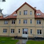Budynek Wspomaganej Społeczności Mieszkaniowej przy ulicy Kętrzyńskiej 29 w Bartoszycach. Do piętrowego budynku prowadzi mokry chodnik, niebo jest szare