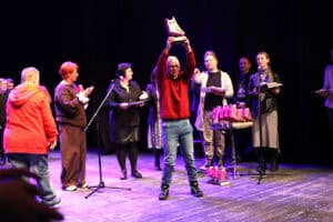 Finałowy moment gali na scenie. W centrum zdjęcia mężczyzna w czerwonym swetrze i jeansach z radosnym uśmiechem unosi wysoko nad głowę czerwoną statuetkę w kształcie sowy. Obok niego, na małym stoliku, ustawiony jest szereg identycznych nagród. W tle widoczna jest grupa osób – organizatorzy i uczestnicy – którzy klaszczą i z uśmiechem obserwują laureata. Po prawej stronie przy mikrofonie stoi kobieta, a po lewej inni uczestnicy w swobodnych strojach. Scena jest jasno oświetlona, co podkreśla uroczysty i pełen entuzjazmu charakter chwili