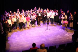 Widok z góry na scenę podczas wielkiego finału wydarzenia. Wszystkie grupy aktorów i wykonawców – łącznie kilkadziesiąt osób – zgromadziły się na deskach teatru. Niektórzy trzymają dyplomy i nagrody. Na pierwszym planie po prawej stronie widać stolik z różowymi statuetkami sów oraz osobę przemawiającą do mikrofonu. W dole kadru widoczne są głowy widzów siedzących w pierwszym rzędzie ciemnej widowni