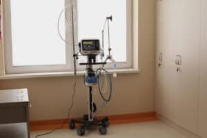 Nowoczesny respirator Hamilton C1-neo na mobilnej podstawie, ustawiony w jasnym pomieszczeniu szpitalnym na tle okna. Urządzenie jest wyposażone w ekran dotykowy z wyświetlonymi parametrami pracy oraz liczne rurki i przewody medyczne. Po prawej stronie znajdują się wysokie, jasne szafy medyczne z kluczykami w zamkach
