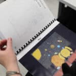 Dłonie osoby trzymają otwartą książkę wydaną w druku powiększonym i brajlu, zbindowaną czarną spiralą. Lewa strona książki zawiera biały arkusz gęsto pokryty wypukłymi punktami systemu Braille’a. Na prawej stronie widnieje barwna ilustracja na ciemnym tle, zatytułowana czarnym drukiem „Kino”. Obrazek przedstawia dwie postacie siedzące tyłem w fotelach kinowych, patrzące na ekran, na którym widać planety i rakietę w przestrzeni kosmicznej. Ilustracja została poddana tyflografii – jej kluczowe elementy mają wyraźną fakturę.