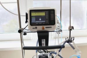 Zbliżenie na panel sterowania respiratora Hamilton C1-neo. Na kolorowym ekranie LCD wyświetlony jest interfejs w języku polskim oraz ustawieniami trybów pracy. Pod ekranem widoczne są oznaczenia producenta oraz naklejka z uśmiechniętym misiem. Po bokach monitora znajdują się fizyczne przyciski sterujące i pokrętło. W tle, za urządzeniem, widoczna jest rama okienna i zarys drzew za szybą