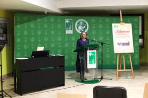 Prof. Iwona Kalinowska w okularach i ciemnoniebieskiej sukience przemawia do mikrofonu przy szklanej mównicy, na tle zielonej ścianki z logo Centrum Onkologii Ziemi Lubelskiej. Obok stoją pianino i plakat wydarzenia „Kolędowanie”.