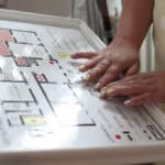 Kobiece dłonie z ozdobnym manicure, dotykiem odczytują plan tyflograficzny parteru budynku. Tablica jest biała, umieszczona w ramie pod kątem ułatwiającym dostęp. Przedstawia czarny, wypukły schemat pomieszczeń (m.in. sklep, kasa, pokój zagadek) z oznaczeniami w brajlu. W dolnej części widoczna jest legenda z wypukłymi symbolami oznaczającymi ściany, okna i pochylnie oraz czerwony punkt z opisem „Tu jesteś”.