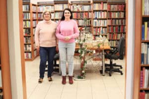 Dwie uśmiechnięte kobiety (bibliotekarki) stojące obok siebie w przejściu między regałami z książkami. Jedna w jasnym golfie i ciemnych spodniach, druga w różowym swetrze i jasnych spodniach. Obok nich dekoracyjna konstrukcja z gałęzi ozdobiona światełkami. W tle widać rzędy regałów z literaturą piękną i popularnonaukową.