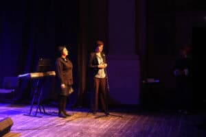 Dwie kobiety na ciemnej scenie podczas oficjalnego wystąpienia. Od prawej Joanna Olszewska w ciemnym żakiecie i wzorzystej bluzce przemawia do mikrofonu na statywie. Po jej prawej stronie stoi Jolanta Grabowska, w czarnej koszuli i okularach. Po lewej stronie znajduje się instrument klawiszowy marki Yamaha na statywie oraz krzesło. Całość oświetlona jest ciepłym, punktowym światłem reflektora