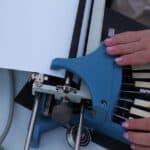 Kobiece dłonie z jasnoróżowym manicure, obsługują maszynę do pisania brajlem. Maszyna ma charakterystyczny niebieski korpus i jest wyposażona w sześć białych klawiszy odpowiadających punktom w komórce brajlowskiej oraz centralny klawisz odstępu. W urządzenie wkręcony jest arkusz grubego, białego papieru, na którym widoczne są już wytłoczone rzędy wypukłych punktów. Lewa ręka spoczywa na górnej części obudowy przy wałku, a prawa operuje na klawiszach. W tle fragment stołu z przyborami, w tym zielona linijka i materiałowe etui.