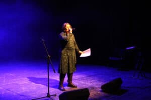 Aleksandra Ziemba w okularach i wzorzystej, zielono-czarnej sukience stoi na środku sceny, śpiewa do mikrofonu trzymanego w dłoni. W drugiej ręce trzyma kartkę z tekstem. Postać jest oświetlona ciepłym światłem punktowym na tle błękitnej poświaty i scenicznego dymu. Obok niej stoi pusty statyw mikrofonowy, a na podłodze widoczne są czarne głośniki odsłuchowe. W tle, po prawej stronie, rysuje się zarys instrumentu klawiszowego