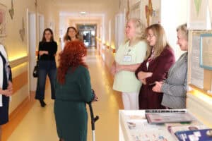 Wnętrze korytarza szpitalnego z dekoracjami w kształcie motyli na ścianach. Cztery kobiety stoją w kręgu, prowadząc rozmowę. Jedna z nich używa kuli łokciowej. Wśród nich kobieta z personelu medycznego w jasnym uniformie i kobiety w strojach biznesowych. W prawym dolnym rogu widoczny fragment biurka z dokumentami i ulotkami