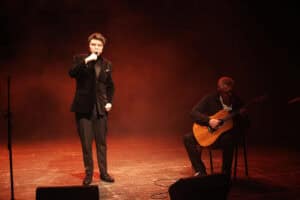 Szymon Woźniak w ciemnym aksamitnym garniturze stoi na scenie i śpiewa do mikrofonu trzymanego w dłoni. Obok niego, na krześle po prawej stronie, siedzi Dariusz Tokarzewski grający na gitarze akustycznej. Scena skąpana jest w ciepłym, pomarańczowo-brązowym świetle i delikatnej mgle. Na pierwszym planie widoczne są fragmenty głośników odsłuchowych, a po lewej stronie stoi pusty statyw mikrofonowy