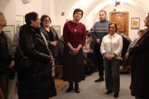 Trzy kobiety stoją w centrum jasnej sali o łukowym sklepieniu, zwracając się do zgromadzonej publiczności. Kobieta w środku (Joanna Olszewska) ma na sobie bordową bluzkę i wzorzystą spódnicę, po jej prawej stronie stoi kobieta w białej koszuli, a po lewej w szarym żakiecie. W tle widoczni są siedzący uczestnicy wydarzenia oraz obrazy zawieszone na ścianach.