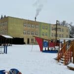 Przedszkole integracyjne w dwupiętrowym prostym budynku w zimowej scenerii, z drewnianym placem zabaw na placu przed budynkiem