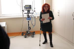 Kobieta o rudych włosach (Renata Polisiakiewicz), w zielonej sukience i czarnych kozakach stoi w jasnym pomieszczeniu medycznym. W jednej ręce trzyma białą teczkę z dokumentami, drugą wspiera się na kuli łokciowej. Obok niej znajduje się nowoczesny respirator marki Hamilton Medical na kołowym stelażu. W tle okno oraz białe szafki medyczne
