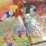 Dwie rozłożone kolorowe książeczki dla dzieci zbindowane metalową spiralą, leżą jedna na drugiej. Strony pokrywa arkusz przezroczystej folii z tekstem wytłoczonym w systemie Braille’a - pismo punktowe pokrywa się z drukiem czarnoocznym i ilustracjami, nie zasłaniając ich. Widoczne są strony z bajek o Złotowłosej oraz o magicznej szafie, z barwnymi rysunkami postaci i tekstem napisanym dużą, czytelną czcionką. Światło odbija się od folii podkreślając wypukłość punktów brajlowskich.