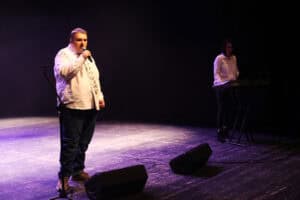 Kamil Dąbrowski w białej koszuli z krawatem w nuty oraz ciemnych spodniach śpiewa do mikrofonu na środku sceny. W tle po prawej stronie, przy instrumencie klawiszowym Yamaha, stoi kobieta w białej bluzce, akompaniująca wykonawcy. Scena jest oświetlona chłodnym, fioletowo-niebieskim światłem z widocznymi na podłodze dwoma czarnymi monitorami odsłuchowymi i statywem mikrofonowym po lewej stronie