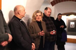 Kobieta z jasnymi włosami, w brązowym kardiganie i czarnym golfie przemawia do zgromadzonych, gestykulując dłońmi. Obok niej, po lewej stronie, stoi mężczyzna w ciemnym garniturze i koloratce, słuchając z uwagą. W tle, w przejściu o ceglanym nadprożu, widoczne są inne osoby uczestniczące w spotkaniu oraz oprawiona grafika na ścianie.