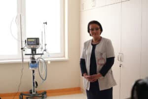 Kobieta w okularach (lek. med. Renata Nieznaj), ubrana w granatową sukienkę i biały fartuch medyczny z identyfikatorem, stoi z rękami splecionymi przed sobą. Po jej prawej stronie widoczny jest respirator Hamilton Medical. Pomieszczenie jest jasne, z rzędem białych szaf w tle i oknem, przez które wpada naturalne światło