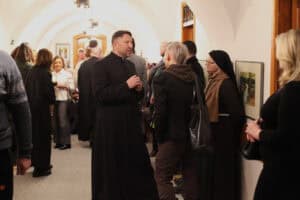 Mężczyzna w czarnej sutannie stoi w centrum jasnego korytarza o łukowym sklepieniu, prowadząc rozmowę z osobą odwróconą tyłem do aparatu. Po prawej stronie, na tle białej ściany z eksponowanymi obrazami, stoi zakonnica w ciemnym habicie i okularach, przysłuchując się dyskusji. W głębi pomieszczenia widoczny jest tłum gości, a po lewej stronie fragment sylwetki mężczyzny w niebieskim, wzorzystym swetrze.