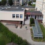 Budynek Warsztatów Terapii Zajęciowej przy SION w Bartoszycach ze schodami i rampą dla osób na wózkach. Kobieta idzie chodnikiem w stronę budynku, w tle osiedle bloków; widok z góry